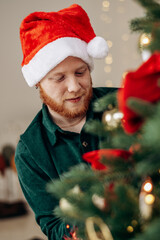 A young red-haired bearded man in the Santa Claus hat is decorating a Christmas tree at home.A New Year's concept.