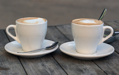 Two white ceramic cups of cappuccino with creamy foam are placed on white saucers, each with a silver spoon resting beside them. The cups sit on a wooden table, creating a cozy and inviting atmosphere