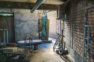Industrial storage area with exposed brick walls, worn concrete, and a bicycle leaning against the wall. Rusted metal pipes, an AC unit, and a wheeled cart contribute to the gritty, urban atmosphere.
