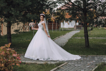 A woman in a white wedding dress is standing in front of a house. She is holding a bouquet of flowers and a veil. The scene is peaceful and serene, with the woman looking happy and content