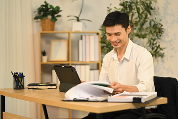Smiling adult businessman analyzing financial reports and using digital tablet at desk