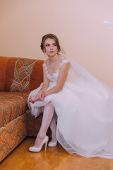 A bride is sitting on a couch, wearing a white dress and white stockings. She is looking at the...