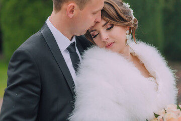 A bride and groom are hugging and kissing each other. The bride is wearing a white fur coat