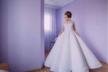 Fototapeta premium A woman in a white dress stands in front of a purple wall. She is wearing a long, flowing gown with a lace collar and a long train. The dress is white with a few small details of lace and embroidery