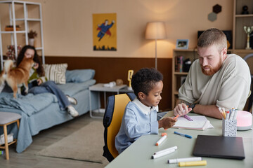 Side view of little Black boy drawing with colorful felt tip pens while supportive dad helping son sitting at desk together in kids room, casual domestic life scene, copy space