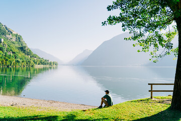 Man sitting at lake meditating with back to camera with calm water and forest in front him, europe