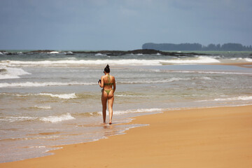 Young tanned slim woman in thong bikini walking by the sand of tropical sea beach, back view. Holidays on sunny coast, concept of freedom, wellness and sun tan
