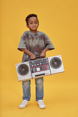 Vertical shot of cute little Black boy smiling at camera holding large boombox playing retro music...