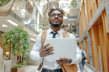 Workplace violence. A stylish professional man holds a laptop, standing confidently in a modern, airy building with green plants and bright wood accents.