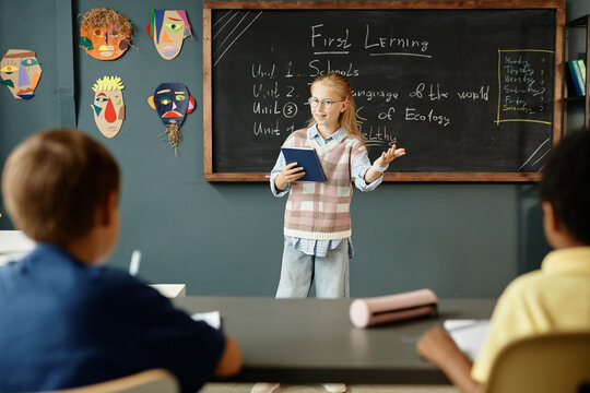 Young intelligent female student answering at blackboard holding notebook, while attentive classmates listening to presentation during lesson in classroom with creative kids crafts on blue wall