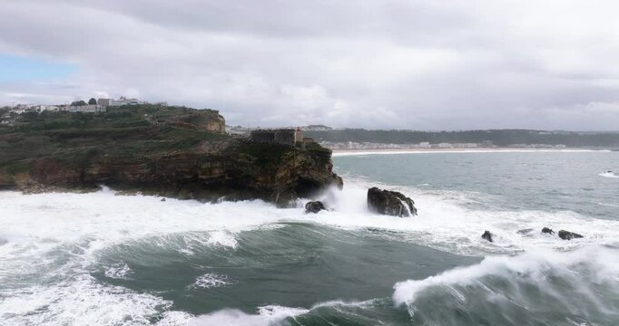 Aerial drone shot of big waves breaking on rocks and cliffs on a day with giant waves in Nazare, Portugal, Europe. Farol da Nazar&eacute; lighthouse visible. Shot in ProRes 422 HQ