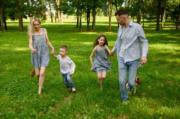 Obraz premium Happy family running and having fun in summer park in nature. Mom, dad and cheerful children, boy and girl, running barefoot on grass, holding hands, dogs running nearby. Parenthood, unity with nature