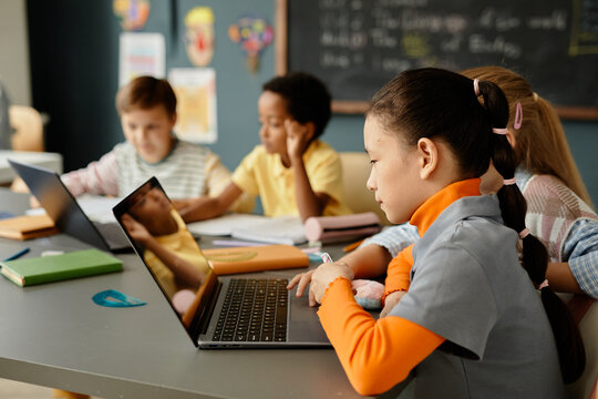 Side view of Central Asian young girl sitting at desk browsing information on laptop fully engaged in class using technology to study at school, copy space