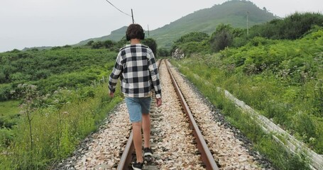 Teenager in plaid shirt and denim shorts walking alone on railway tracks in a green countryside, captured from the back with themes of solitude and introspective journey