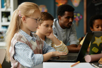 Side view of smart young blonde girl in glasses studying at table using laptop during lesson in modern classroom, copy space