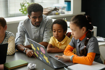 African American male teacher helping younger multiethnic students with using laptop explaining...