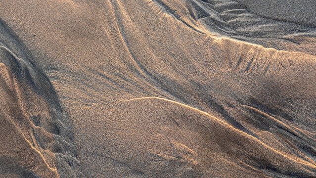 Natural patterns in the beach sand created by wind and waves, highlighted by soft, warm sunlight, form intricate lines and textures in a serene coastal setting.