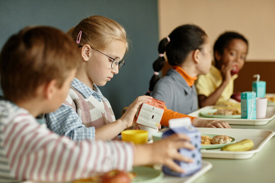 Side view of young girl pouring fresh juice or milk into cup sitting at table with classmates while eating lunch in school cafeteria, copy space