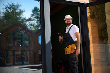 Construction worker installing door in a new house construction site. Man in working attire install front door. Builder in uniform with screwdriver adjusting front glass door in new beautiful house.