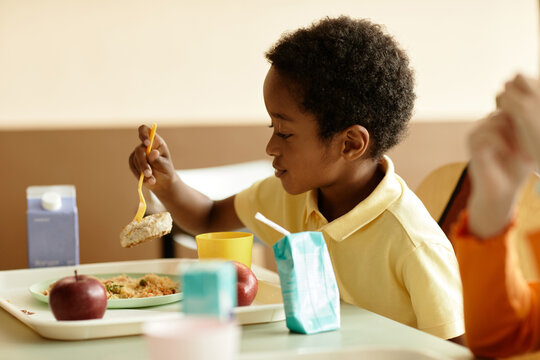 Side view of smiling African American boy sitting at table enjoying eating lunch in school canteen, copy space