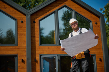 Architect hold plan. Bearded architect wearing hardhat holding project blueprints paper plans standing near comlex of new modern houses looking to something at construction site.