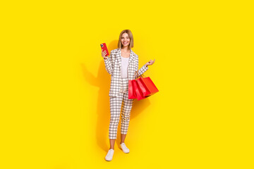 Confident businesswoman with shopping bags and smartphone, dressed in white jacket against yellow backdrop