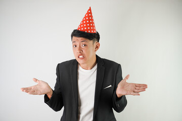 Fierce young asian man wearing red party hat, white shirt and black suit with complaining gesture,...