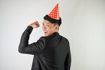 Energetic young asian man wearing red party hat, white shirt and black suit is flexing his bicep muscle, isolated over white background. Concept for Christmas Holiday and New Year Party