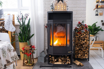 Metall black stove, burning hearth fireplace in white Festive interior of house is decorated for Christmas and New Year, Christmas tree. firewood in the woodpile, cozy and heating of home