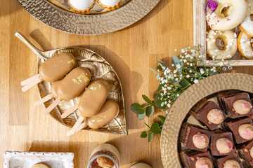 An assortment of refined desserts displayed on a wooden table. Gold-dusted caramel ice cream bars, chocolate mousse squares, and delicately decorated white-frosted donuts add an elegant touch, ideal f