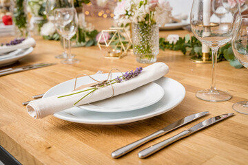A rustic yet refined table setting featuring a white plate with a lavender sprig-tied napkin, polished silverware, and glassware. Floral arrangements and a geometric gold candle holder add elegance to