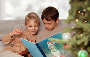 Two happy brothers sitting on the sofa and reading a book by Christmas tree. Kids play at home.