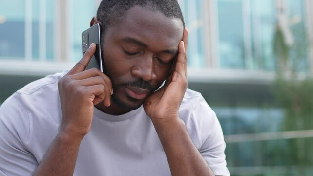 Sad tired lonely disappointed African man alone talking on smartphone on city street outdoor. Unhappy person making answering call by cellphone experiencing grief getting bad news. Stressed man