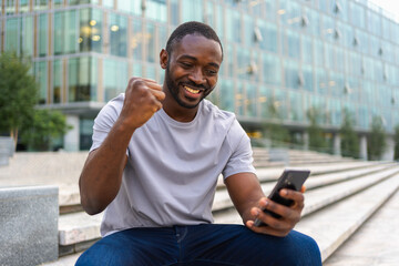 Happy African American man euphoric winner with smartphone on street in city. Person guy looking at cell phone reading great news getting good result winning online bid feeling amazed. Winning gesture