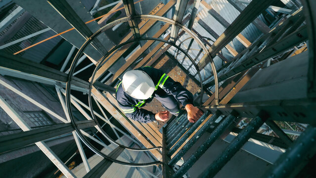 Worker Climbing Industrial Ladder with Safety Gear. Top view of a worker in a hard hat and reflective gear climbing a caged industrial ladder.