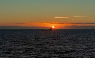 Fototapeta premium Silhouette of a transport ship close to the sun. Wind farm in the background, North Sea during sunset, Netherlands.