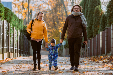 Fototapeta premium A loving couple strolls through a sunlit park with their young son, surrounded by the vibrant colors of autumn, enjoying a joyful and peaceful family moment together.