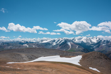Aerial top view to stony pass and snow-white glacier on rocky hill in sunlight among high mountains in sunny day. Large colorful mountain range with snowy peaks in far away under clouds in blue sky.