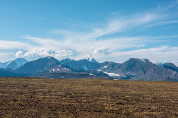 Top view from sunlit grassy stony plain of pass to large rocky mountain range with glacier and high snow pointy peak in bright sun under clouds in blue sky. Big snowy peaked top far away in sunny day.