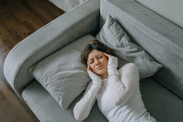 Young woman suffering from strong headache lying on sofa at home, touching temples with fingers,...