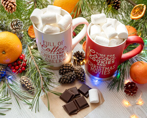 Festive Christmas scene with two mugs of hot chocolate topped with marshmallows, surrounded by pine branches, pinecones, oranges, chocolate, and holiday decorations, creating a cozy holiday atmosphere