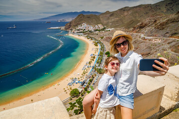 A mother and daughter, both wearing sunglasses, share a joyful moment with the scenic Las Teresitas...