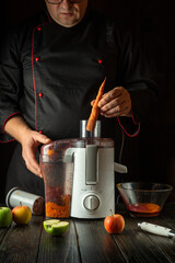 Chef manually adding raw carrots to electric juicer. Making healthy vegetable juice on hotel kitchen counter for breakfast