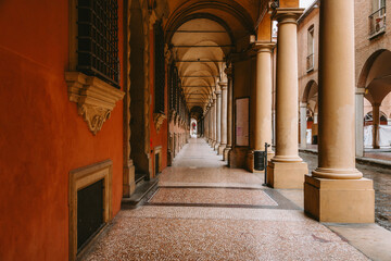 Warm sunlight illuminates the long, empty colonnade with its repetitive arches and columns, creating a striking perspective and highlighting the architectural beauty of bologna