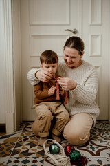 Grandmother and grandson sit on the floor in the kitchen and tie ribbons on Christmas  glass vintage balls.