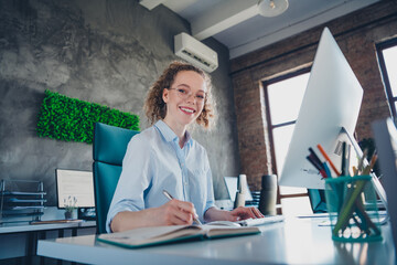 Confident young businesswoman in formal shirt sitting at workstation and writing notes in modern...
