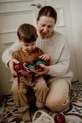 Grandmother and grandson are sitting on the floor in the kitchen and unwrapping Christmas tree glass vintage balls.