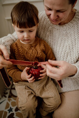 Grandmother and grandson in knitted sweaters tie a velvet ribbon on a red vintage Christmas tree ball. Creating a Christmas decoration