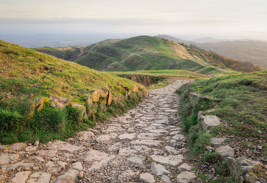 An ancient Stone Age fort in the Malvern Hills UK, with landscape in the distance. A stone path in the foreground leads the eye to mid ground showing pathways cut into the side of a hill. 