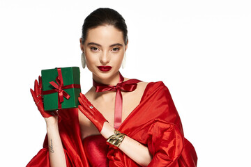 A stunning woman poses elegantly, showcasing a stylish red outfit while holding a green gift box.